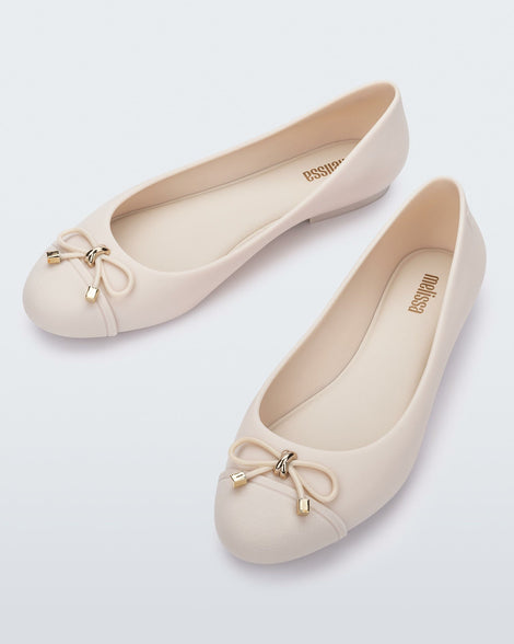 An angled front and side view of a pair of light beige Melissa flats with a beige bow detail with gold accents on the toe
