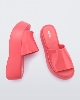 Side view and top view of a pair of Melissa  Becky platfrom slides in bright pink colorway. Melissa logo is printed in white on the insole of the shoe. 