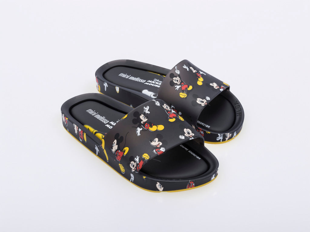 An angled front view of a pair of black Mini Melissa Beach slides with a black base and a pattern of Disney's Mickey Mouse in different poses.
