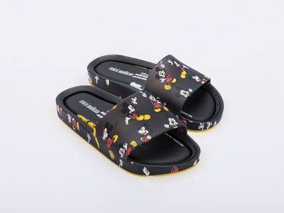 An angled front view of a pair of black Mini Melissa Beach slides with a black base and a pattern of Disney's Mickey Mouse in different poses.