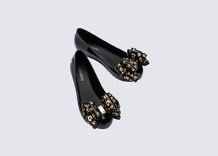 Top view of a pair of black Ultragirl Sweet X kids flats with a bow that has chrome flowers on it