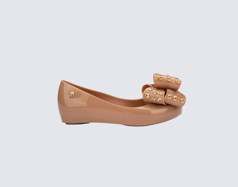 Side view of a beige Ultragirl Sweet X kids flats with a bow that has chrome flowers on it