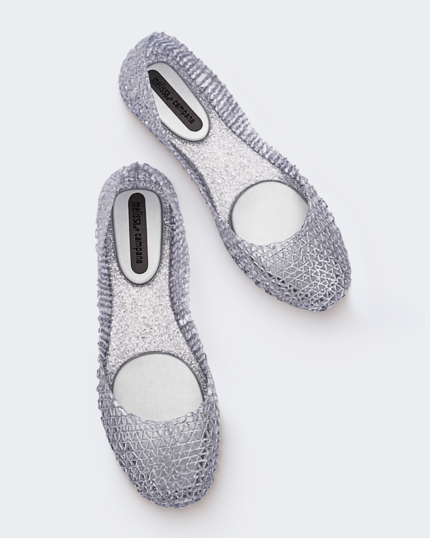 An angled top view of a pair of glitter silver Melissa Campana flats with a distinctive interwoven pattern