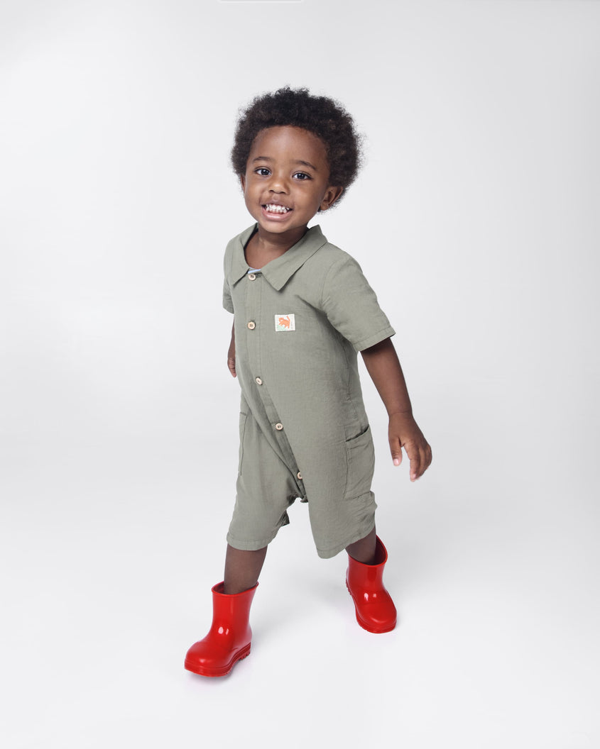 A young child model in a gray one piece outfit wearing a pair of red Melissa Welly boots.