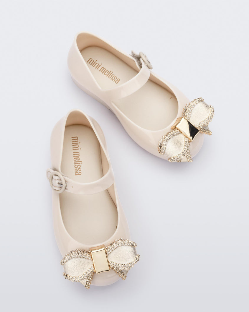 Top view of a pair of beige Mini Melissa Sweet Love baby mary jane flats with a metallic gold bow.