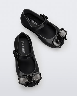 Top view of a pair of black Mini Melissa Sweet Love baby mary jane flats with a metallic gray bow.