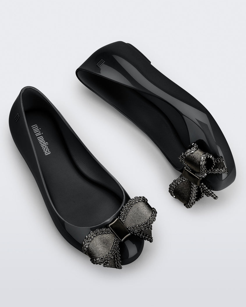 Side and angled view of a pair of black Mini Melissa Sweet Love kids flats with metallic gray bow.