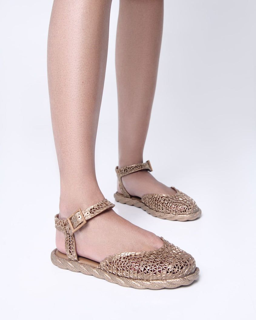 Model's legs wearing a pair of gold Melissa Papel Espadrille Campana sandals with braided sole detail and ankle strap