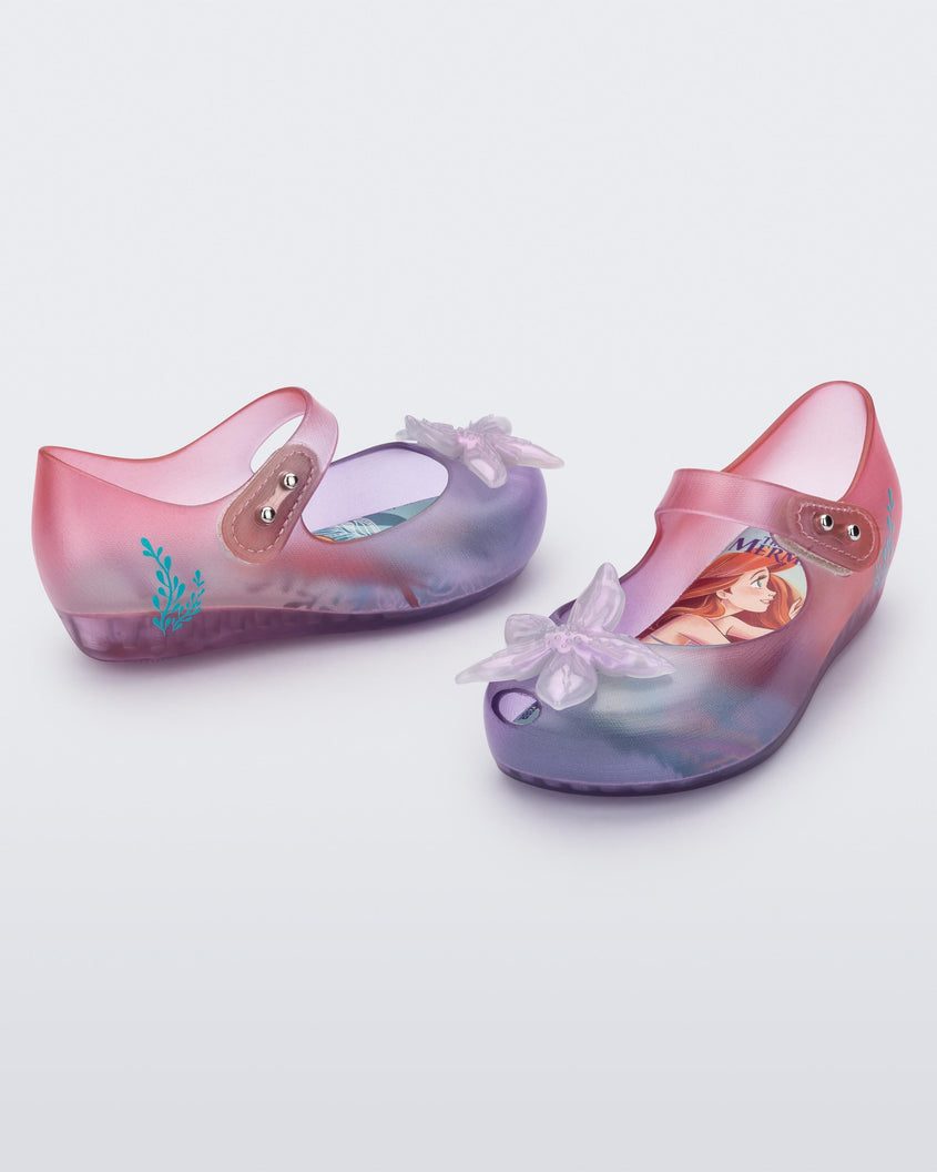 Angled view of a pair of transparent pink and purple Mini Melissa Ultragirl + Disney Little Mermaid flats with a pearly white flower on top