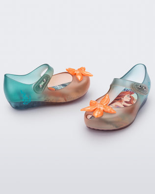 Angled view of a pair of transparent green and orange Mini Melissa Ultragirl + Disney Little Mermaid flats with a pearly orange flower on top