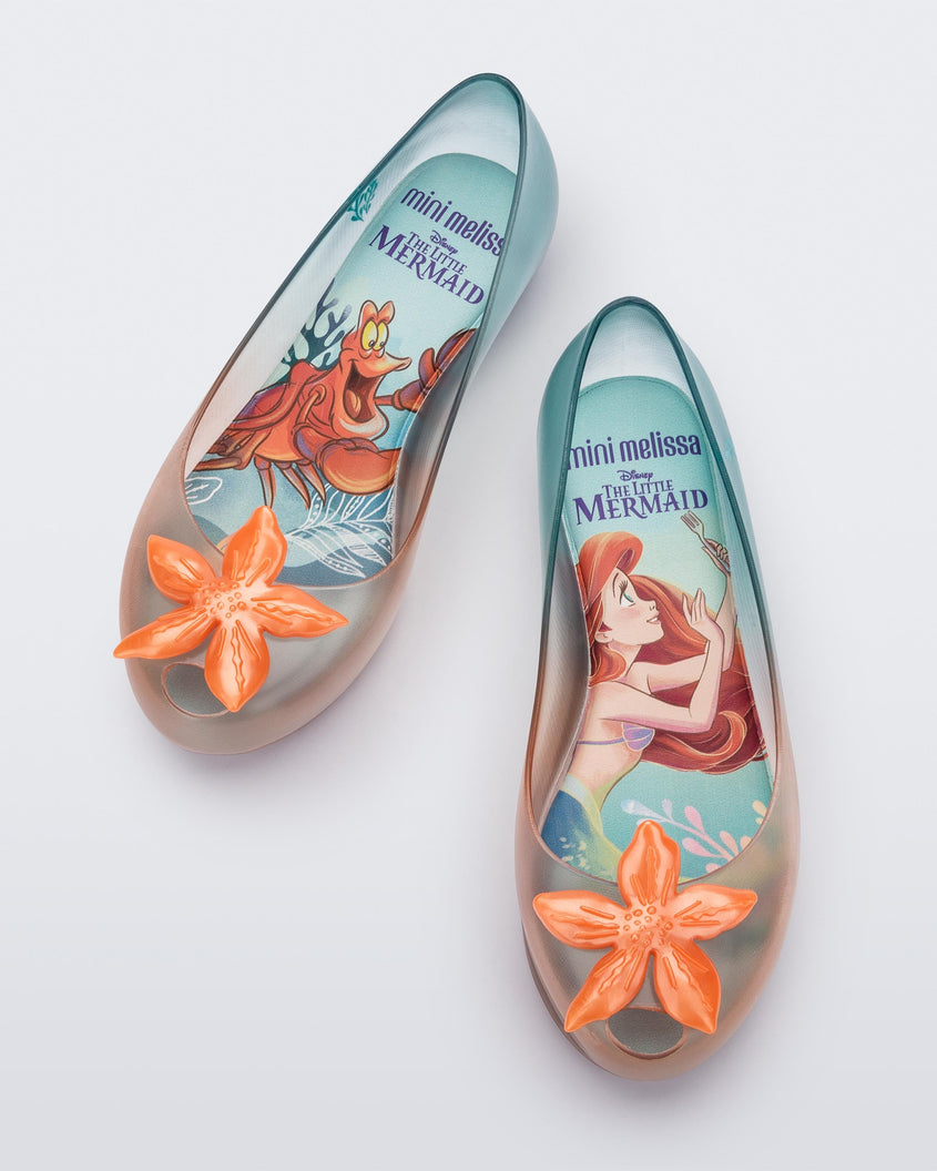 Top view of a pair of transparent green and orange Mini Melissa Ultragirl + Disney Little Mermaid flats with a pearly orange flower on top
