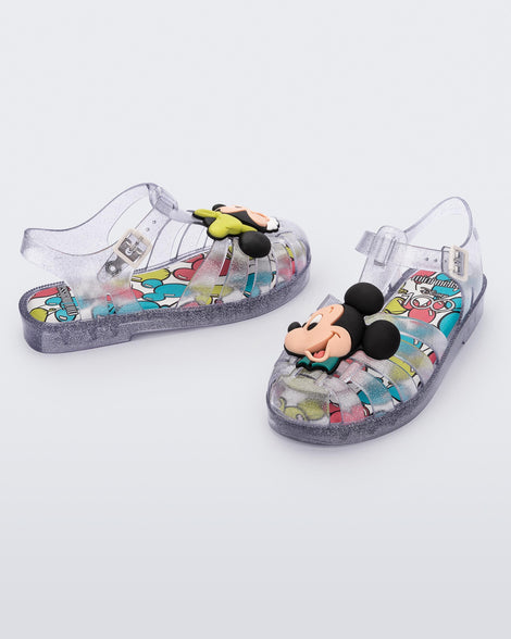 Angled view of a pair of clear glitter Mini Melissa Possession + Disney 100 sandal with Mickey on one foot and Minnie on another foot.