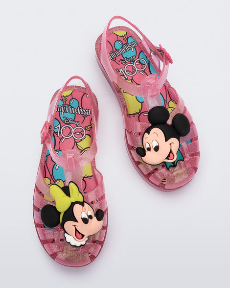 Top view of a pair of clear pink glitter Mini Melissa Possession + Disney 100 sandal with Mickey on one foot and Minnie on another foot.