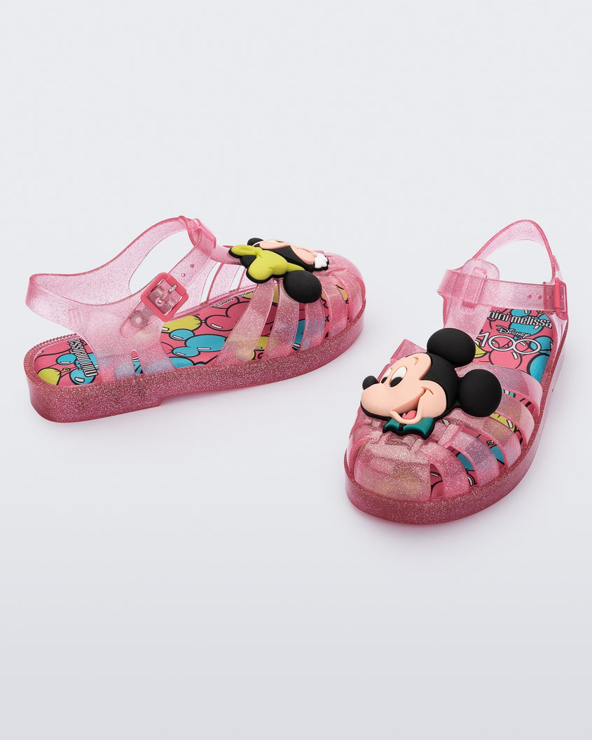 Angled view of a pair of clear pink glitter Mini Melissa Possession + Disney 100 sandal with Mickey on one foot and Minnie on another foot.
