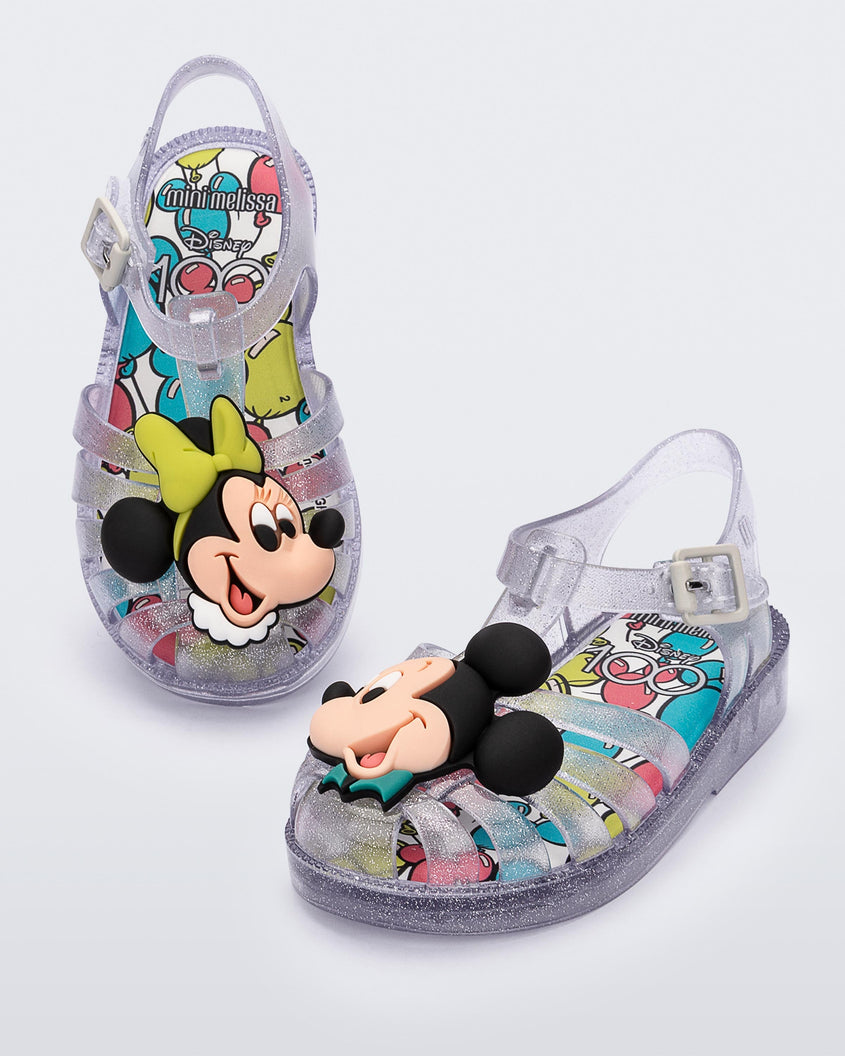 Angled view of a pair of clear glitter Mini Melissa Possession + Disney 100 sandal with Mickey on one foot and Minnie on another foot.