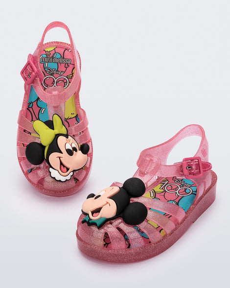 Angled view of a pair of clear pink glitter Mini Melissa Possession + Disney 100 sandal with Mickey on one foot and Minnie on another foot.