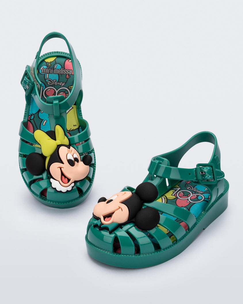 Angled view of a pair of green Mini Melissa Possession + Disney 100 sandal with Mickey on one foot and Minnie on another foot.