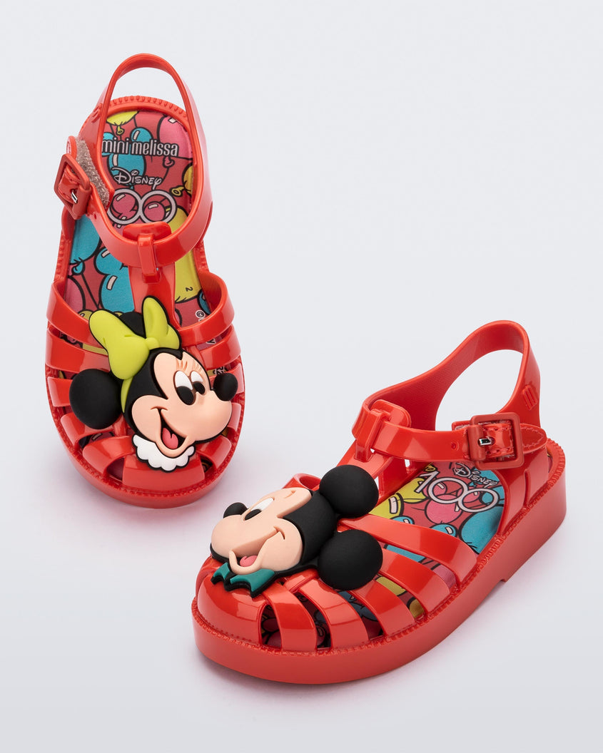 Angled view of a pair of red Mini Melissa Possession + Disney 100 sandal with Mickey on one foot and Minnie on another foot.