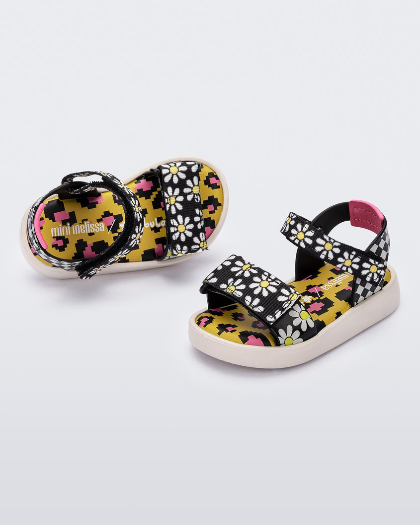 Angled view of a pair of white, black and yellow patterned Mini Melissa Pula Pula + Fabula sandals.