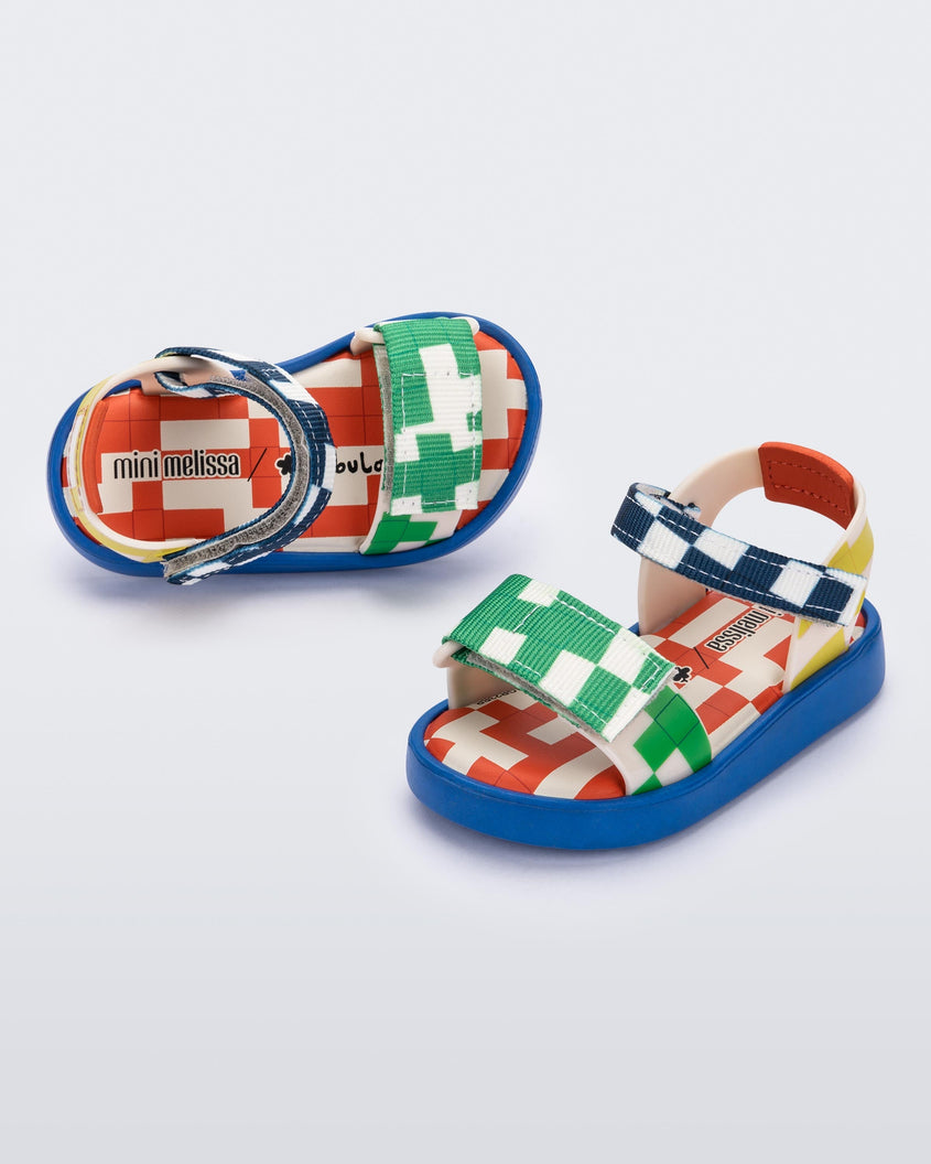 Angled view of a pair of  blue, yellow, green and beige patterned Mini Melissa Pula Pula + Fabula sandals.