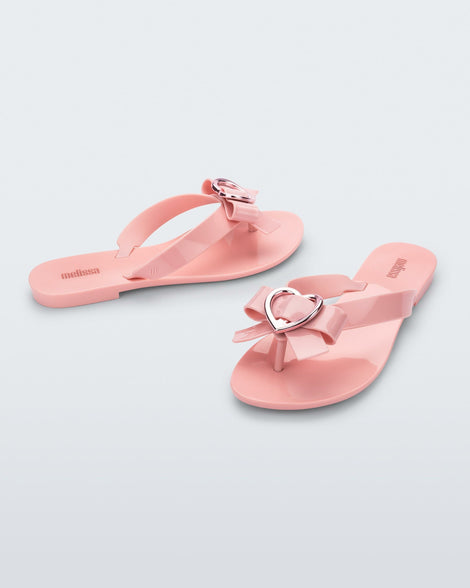 An angled front and side vie of a pair of pink Melissa Harmonic Heart flip flops with a pink bow and metallic pink heart detail on the straps