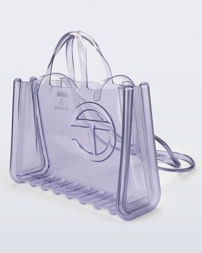 Angled view of a clear Melissa Large Jelly Shopper + Telfar bag with handles and a body strap