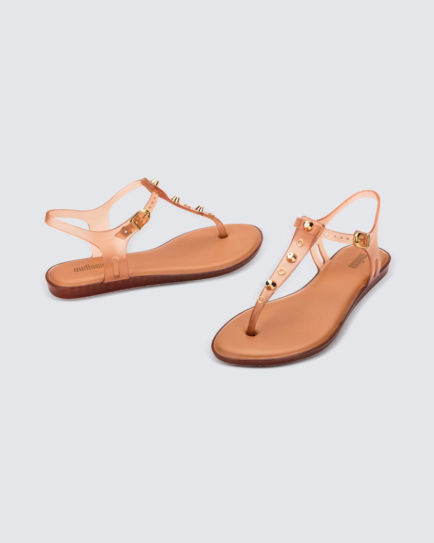 Angled and side view of a pair of clear beige Melissa Solar studs sandals with gold studded t-strap and gold buckle.