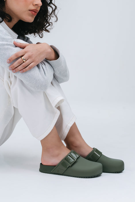 Model in a grey sweater and white pants wearing a pair of green Cozy clogs