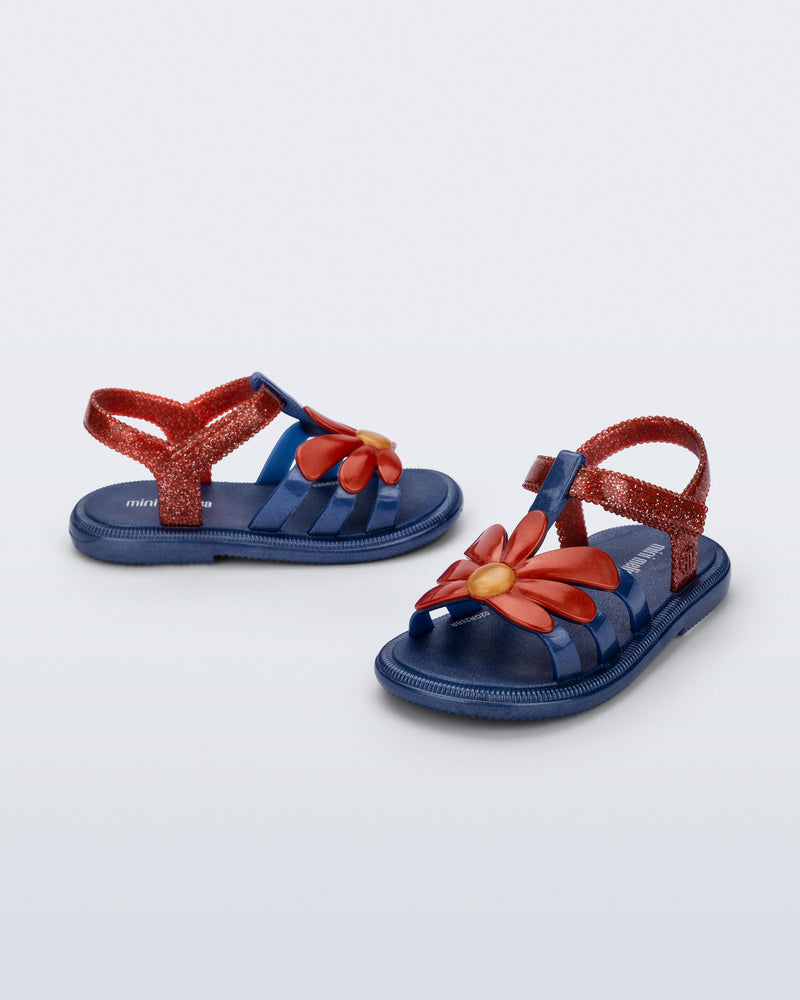 Side and angled view of a pair of glitter blue Hip Bloomy baby sandals with red daisy on top and red straps