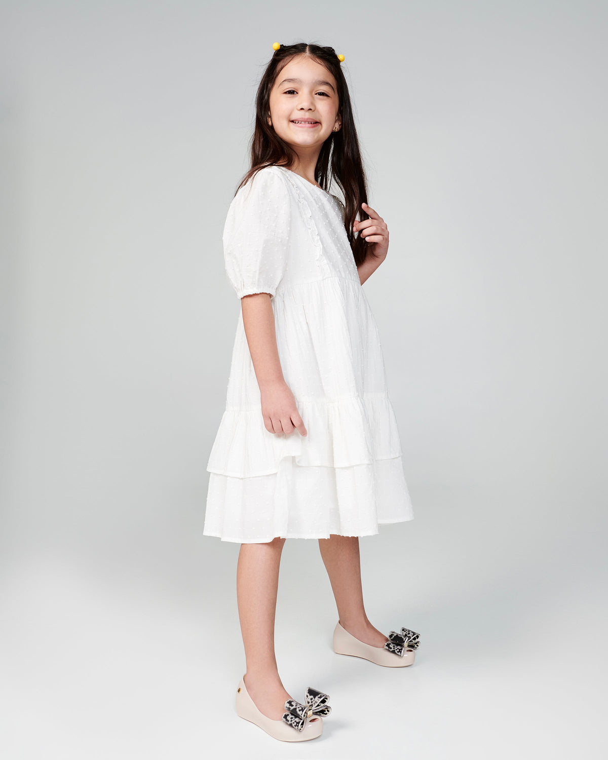 Child model in a white dress wearing a pair of beige kids Ultragirl Classic Bow kids flats with black and clear bow