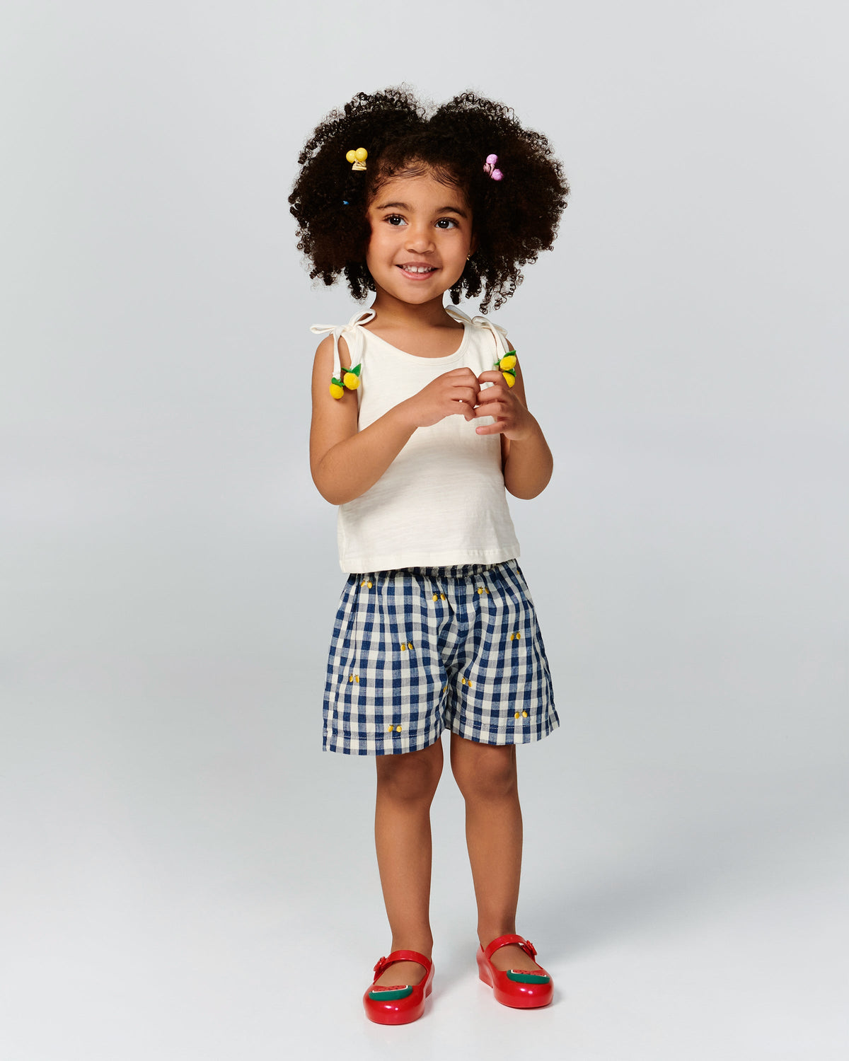 Child model in a white shirt and black and white shorts wearing a pair of red Sweet Love Picnic baby flats with watermelon slices
