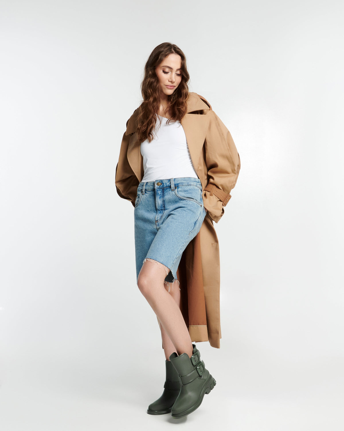 Model in jean shorts, white shirt and camel colored coat wearing a pair of green Bang boots with side buckles