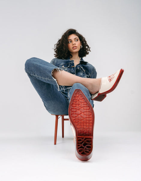 Model in cut off blue jeans and jacket sitting in a chair wearing a pair of beige with red heel and accents Texas Mule heels and showing the bottom sole of one.