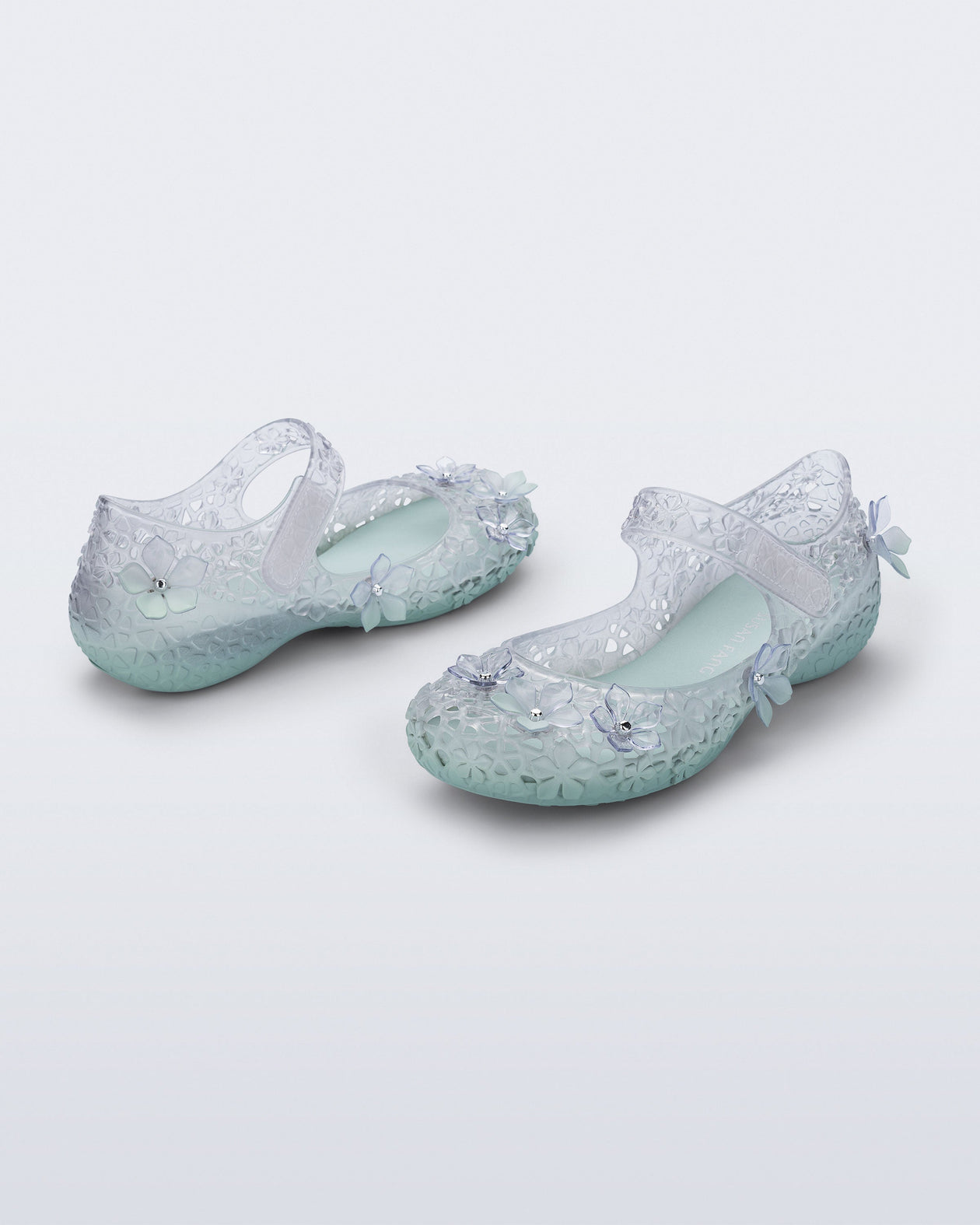 Back and side angled view of a pair of clear Melissa + Susan Fang Luna Bloom flats with flower adornments and misty green sole