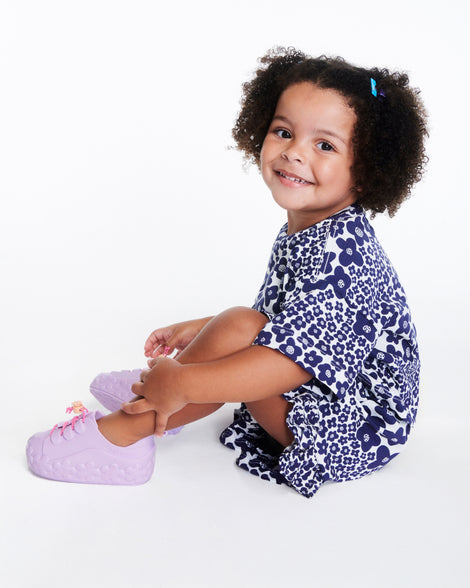child model in a navy dress wearing a pair of purple Bloomy Sneakers with pink laces and flower details on the sole