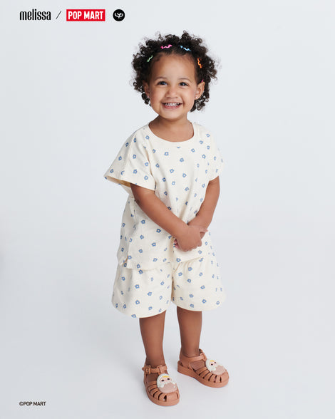 Child model in a white with blue shapes short set wearing a pair of beige Possession + Pop Mart Pucky baby fisherman sandals