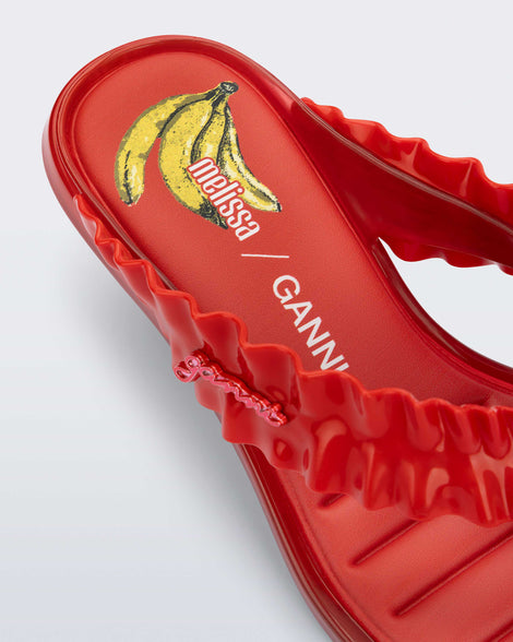 Close up view of the heel of a  red Flip Flop Kitten Heel + GANNI sandal with banana print foot bed and faux ruffle strap