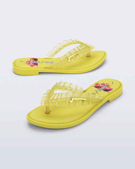 Side and angled view of a pair of yellow Flip Flop Slim + GANNI sandals with a cherry print foot bed and faux ruffle strap