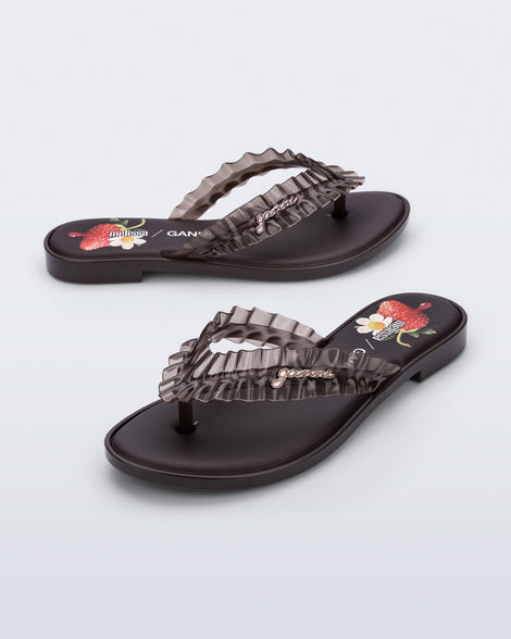 Side and angled view of a pair of  brown Flip Flop Slim + GANNI sandals with a strawberry print foot bed and faux ruffle strap