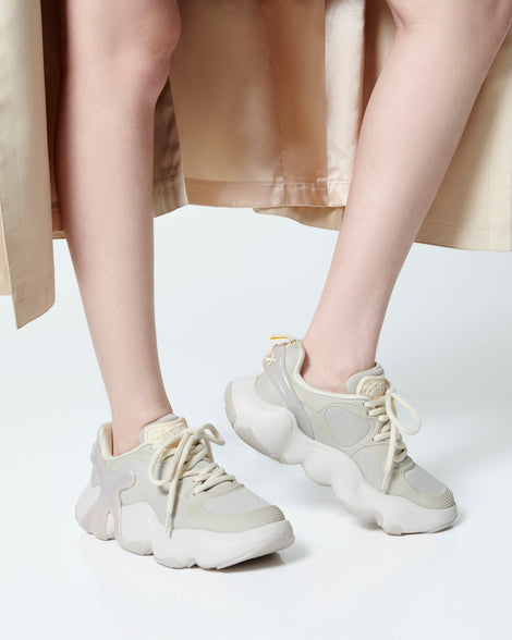 Model's legs in a beige trench coat wearing a pair of beige Melissa X Rush sneakers