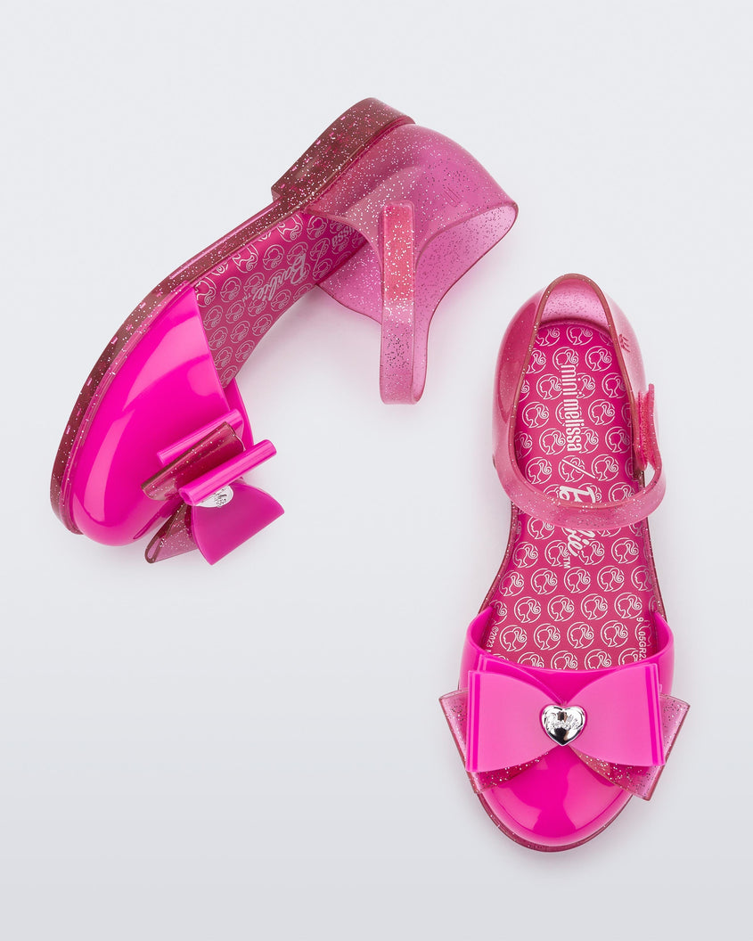 A top and side view of a pair of glitter pink Mini Melissa sandals with a Barbie bow detail on the front toe, pink glitter ankle strap and a Barbie logo sole