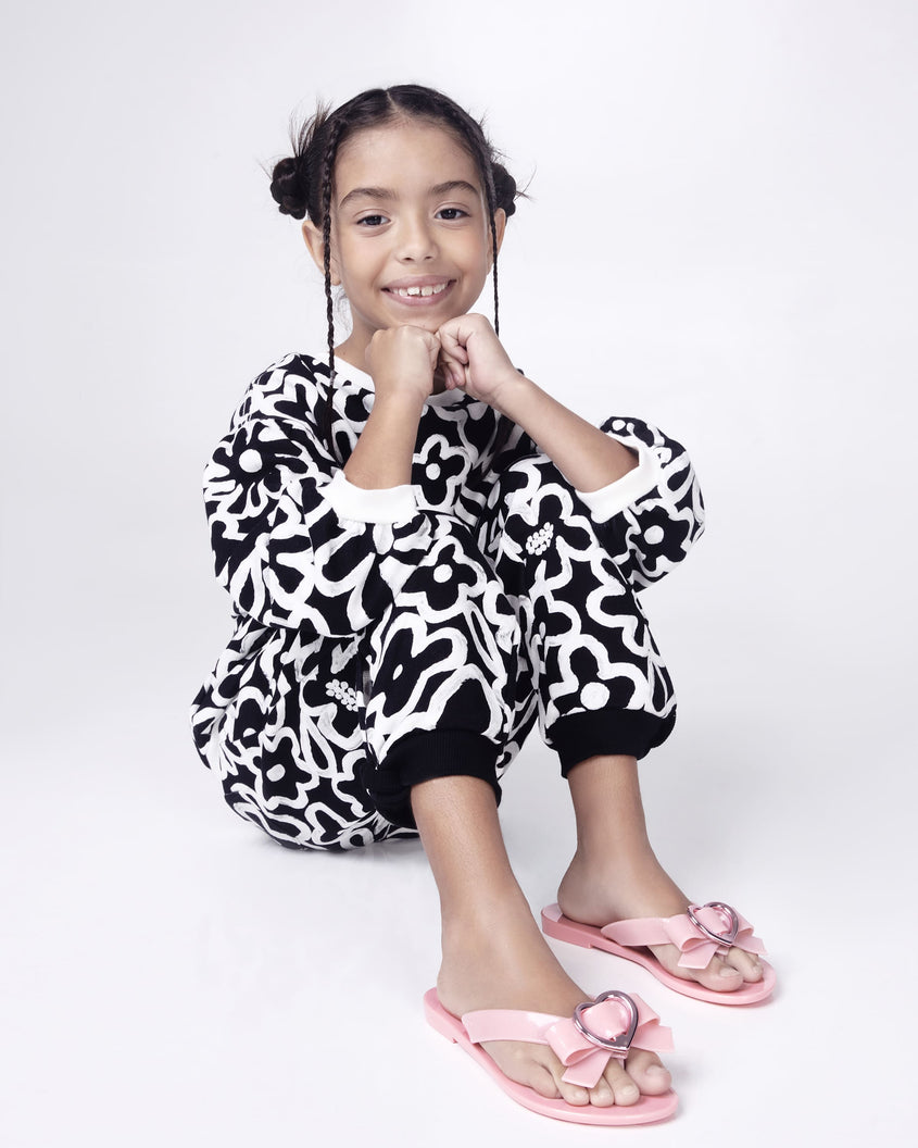 A kid model posing for a picture in a patterned top and bottom, wearing a pair of pink Mini Melissa Harmonic Heart flip flops with a pink bow and metallic pink heart detail on the straps