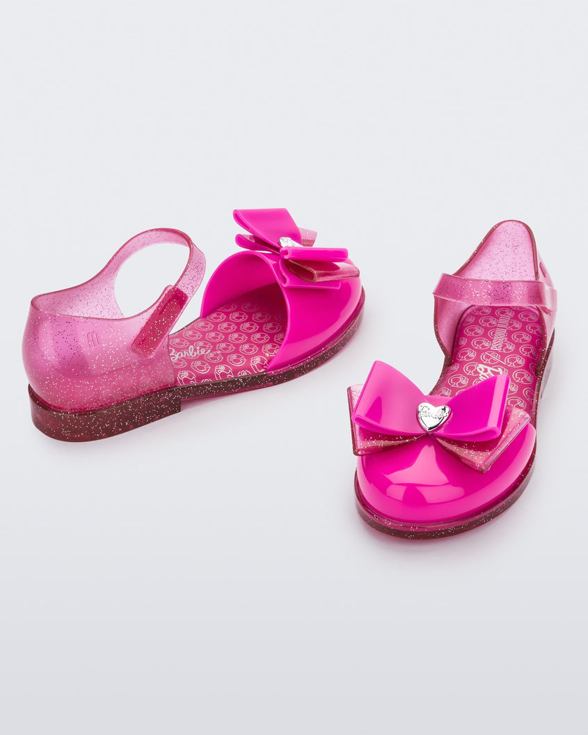 An angled front and side view of a pair of glitter pink Mini Melissa sandals with a Barbie bow detail on the front toe, pink glitter ankle strap and a Barbie logo sole
