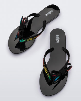 Top view of a pair of black Harmonic M Lover adult flip flop with rainbow Melissa logo on strap