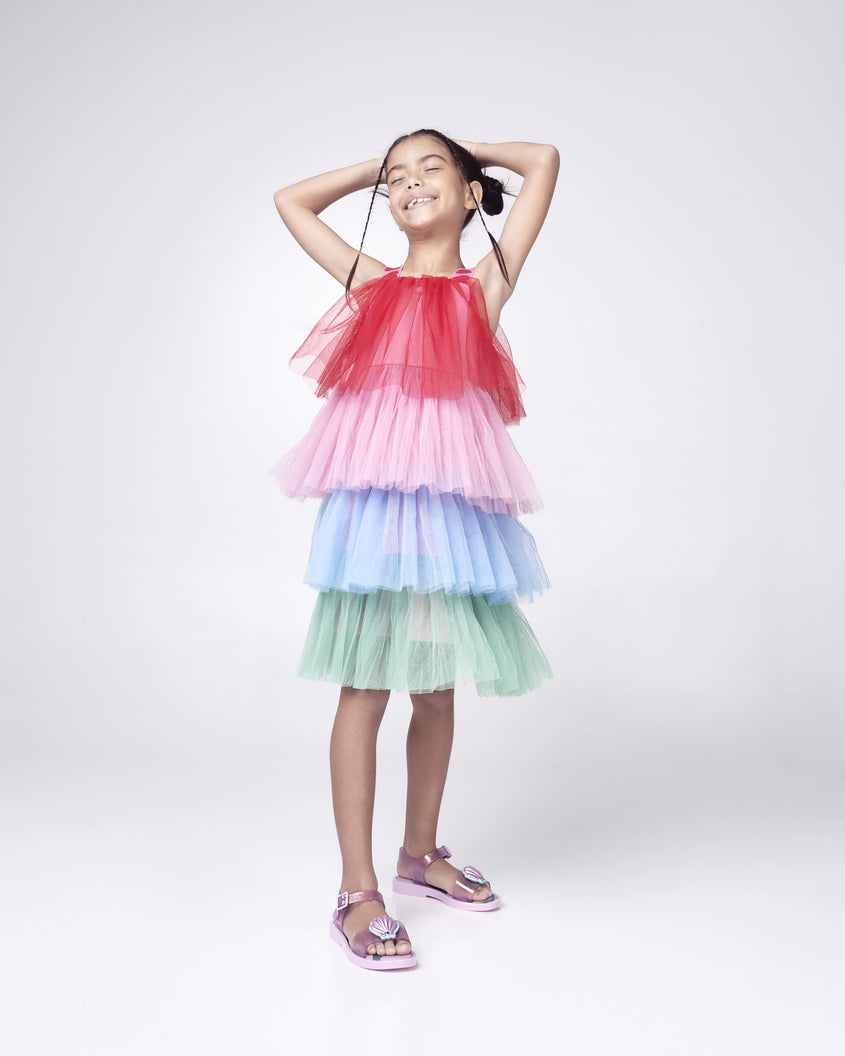 A kid model posing for a picture in a multicolor dress and a pair of glitter pink Mini Melissa Mar Sandal Princess sandals with a seashell detail on the front strap, an ankle strap and Princess Ariel soul