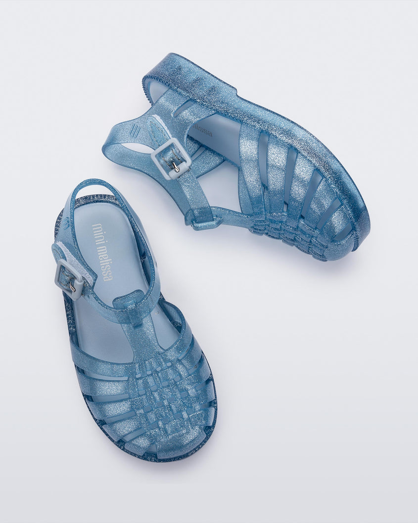 Top and side view of a pair of Mini Melissa Possession baby sandals in glitter blue with velcro buckle closure on the ankle straps