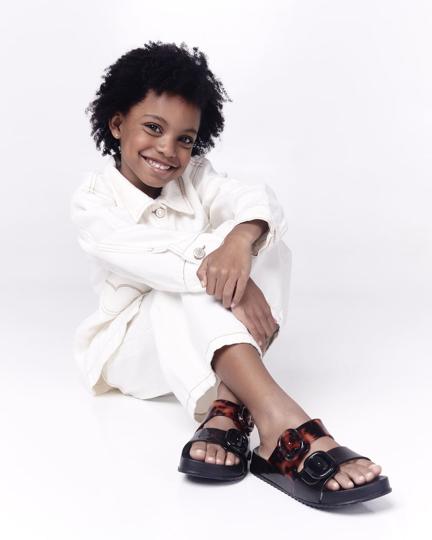 A kid model posing for a picture in a white top and bottom, wearing a pair of black tortoiseshell Mini Melissa Cozy slides with two front straps with buckle details
