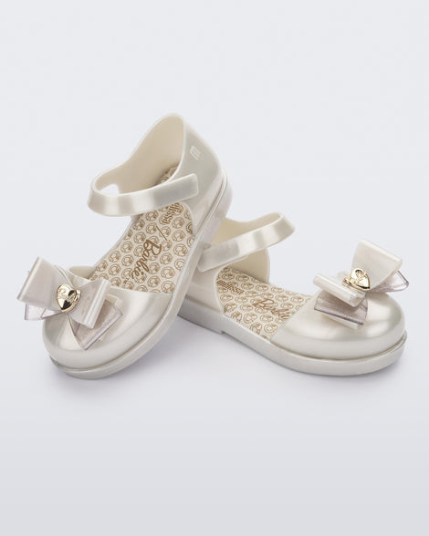 An angled top and side view of a pair of white metallic Mini Melissa sandals with a Barbie bow detail on the front toe, white metallic ankle strap and a Barbie logo sole