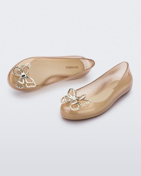 An angled front and top view of a pair of glitter beige Mini Melissa Sweet Love Butterfly flats with a gold butterfly detail on the toe
