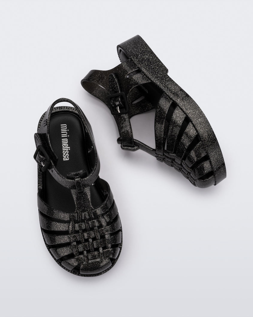 Top and side view of a pair of Mini Melissa Possession baby sandals in glitter black with velcro buckle closure on the ankle straps